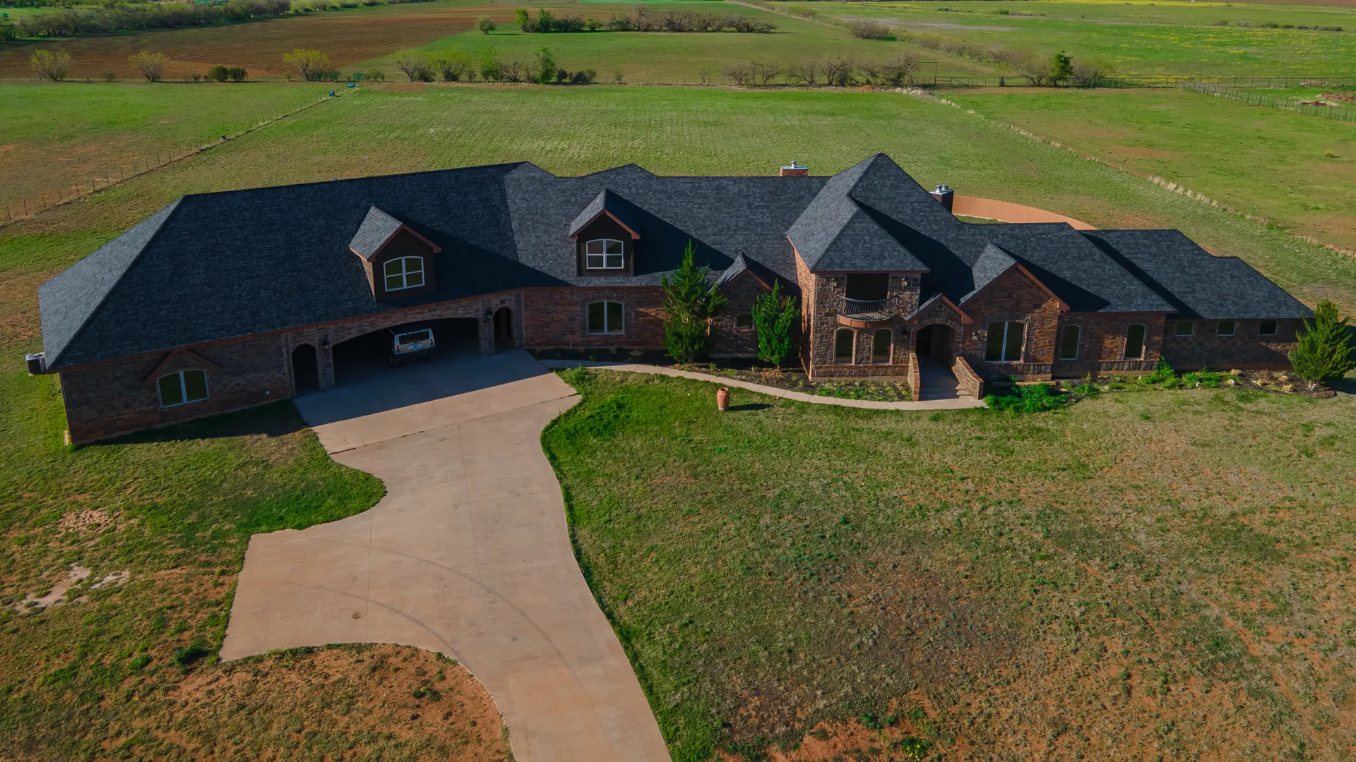 Large brick and stone home with a dark shingle roof, seen from above, with open fields behind it