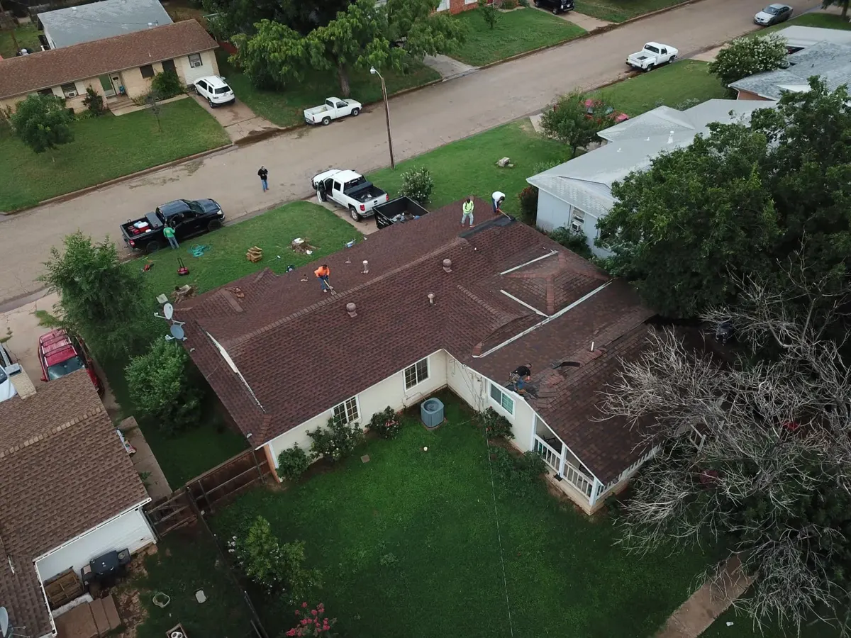 Drone view of a crew working on a house roof, with work trucks in the street and a dumpster by the house